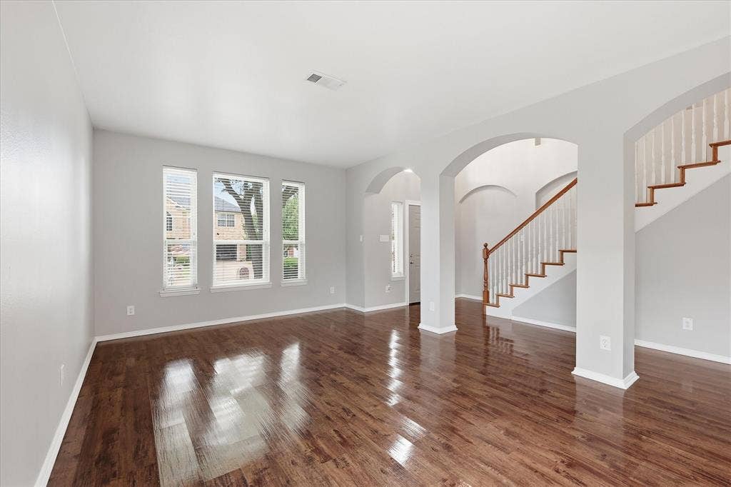 Expansive room featuring rich wood-finish flooring, multiple windows with blinds, a grand staircase with wood handrails, and arched doorways