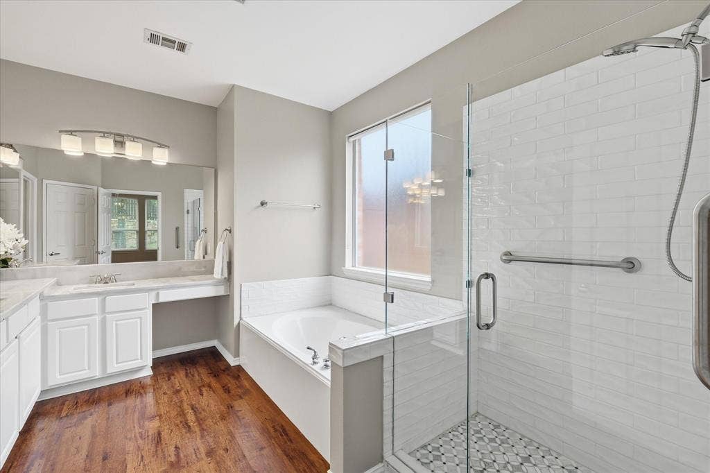 Wood-finish flooring, white vanity with dual sinks, built-in soaking tub, glass-enclosed shower with subway tile, and frosted window