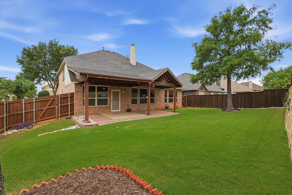 Expansive backyard featuring a manicured lawn, full privacy fence, mature trees, and a brick patio with an attached covered outdoor living area