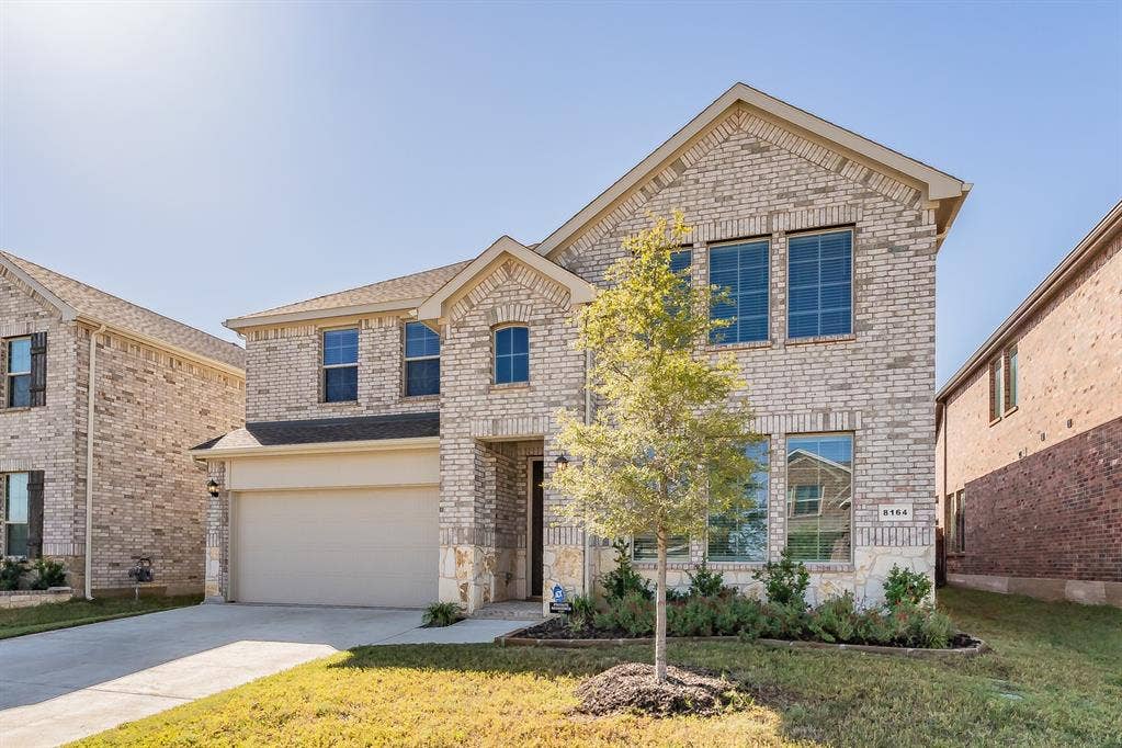 Two-story residence featuring a light-colored brick facade, stone accents, an attached two-car garage, multiple windows, and a newly landscaped front yard