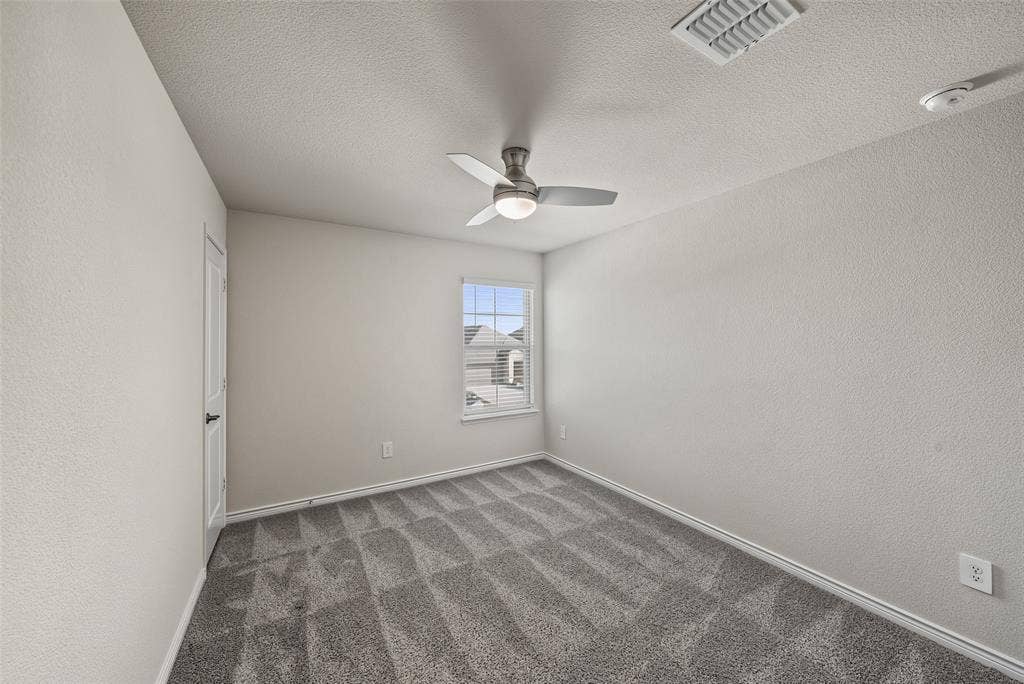 Textured neutral walls, gray carpeting, single window with white trim, white door with dark hardware, and a ceiling fan with integrated lighting