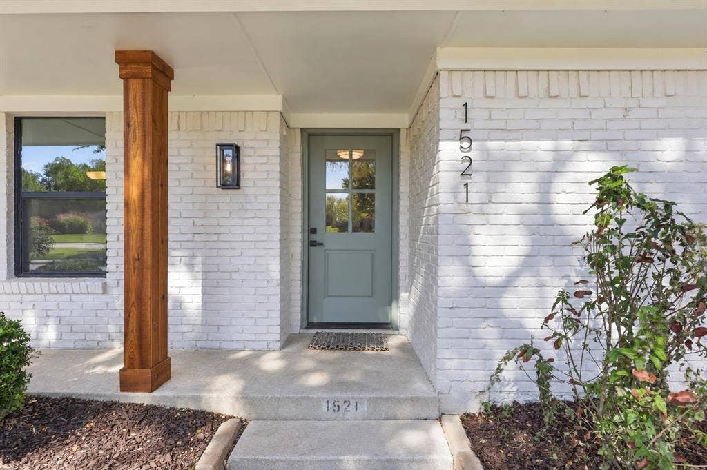 1521 Glenwick Drive, Plano, TX 75075 Exterior featuring a painted brick facade, covered entryway with a wood-finished pillar, four-panel glass-inset entry door, and dark-framed wall-mounted lighting