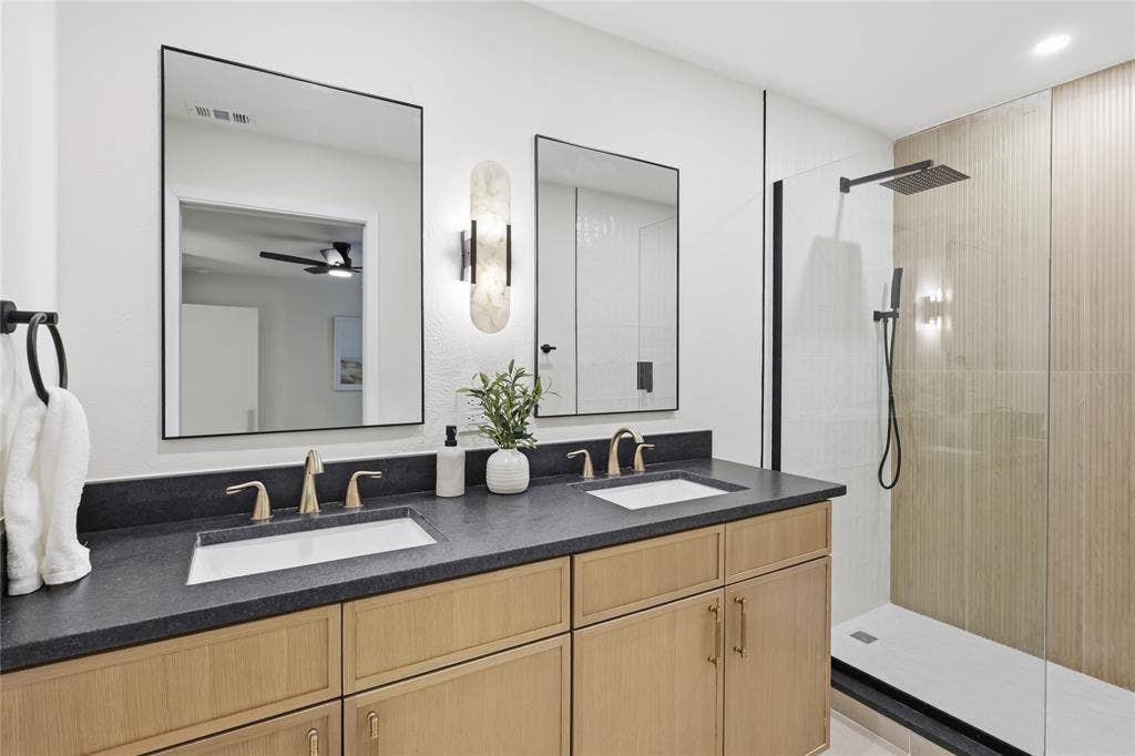 1521 Glenwick Drive, Plano, TX 75075 Double vanity featuring wood-finish cabinetry, dark solid-surface countertops, rectangular undermount sinks, gold-tone faucets, and two frameless mirrors with a vertical wall-mounted light fixture