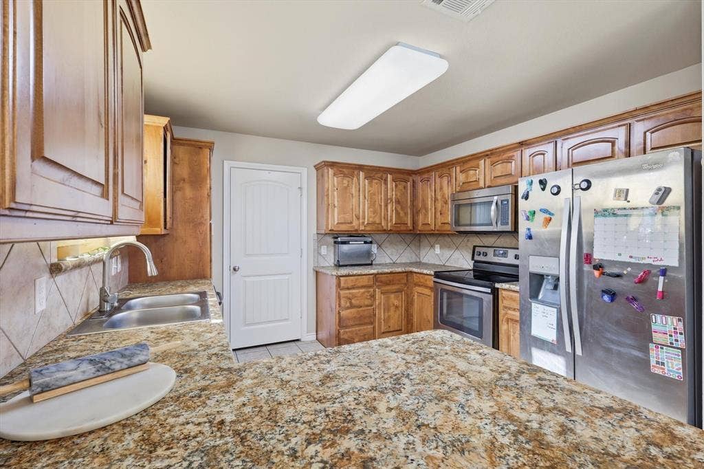 Kitchen featuring wood cabinetry, stainless steel appliances, granite-style countertops, tile backsplash, and a double basin sink