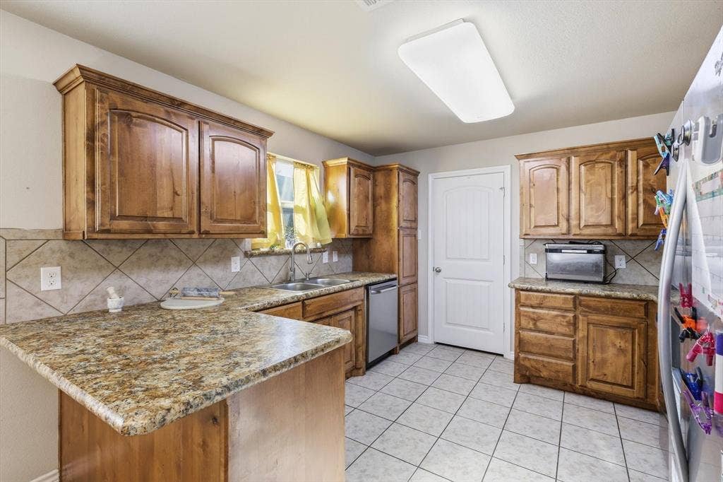 Warm wood cabinetry, granite-style countertops, tile backsplash, stainless steel dishwasher, and tiled flooring