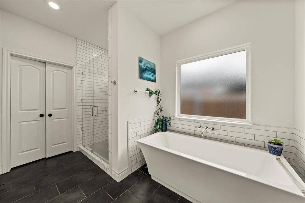 Freestanding soaking tub with wall-mounted faucet, glass-enclosed shower with subway tile surround, and dark tile flooring