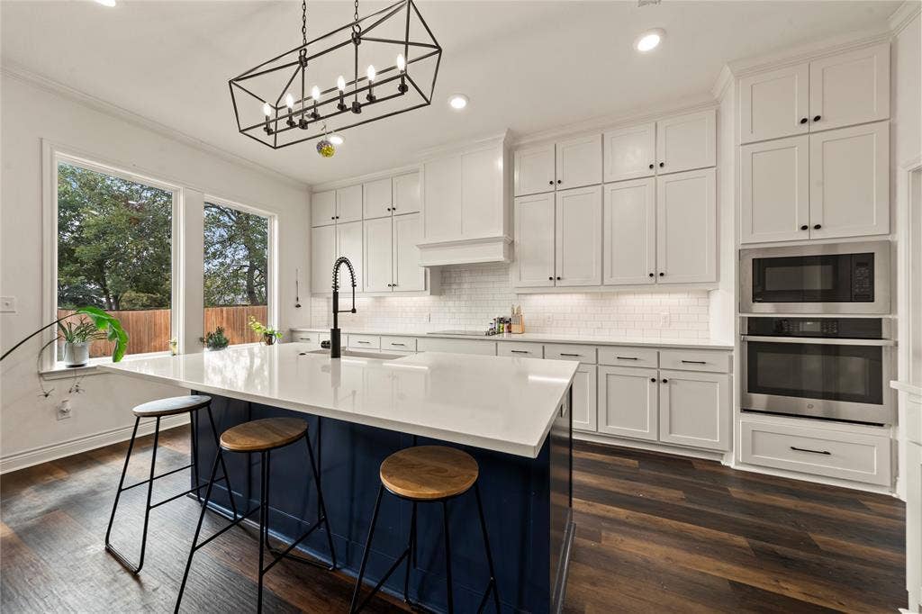 Modern kitchen featuring white shaker cabinetry, dark wood-finish flooring, a central island with white countertop, stainless steel double ovens, and a black open-frame chandelier