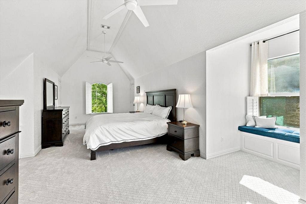 Vaulted ceiling with dual ceiling fans, arched window with plantation shutters, window seat with built-in storage, and wall-to-wall carpeting