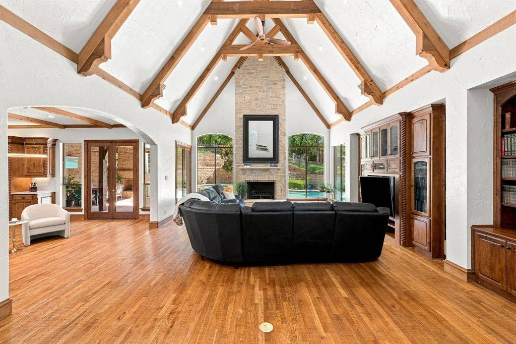 Vaulted ceiling with exposed wood beams, stone-faced fireplace, hardwood flooring, built-in entertainment center, and integrated shelving