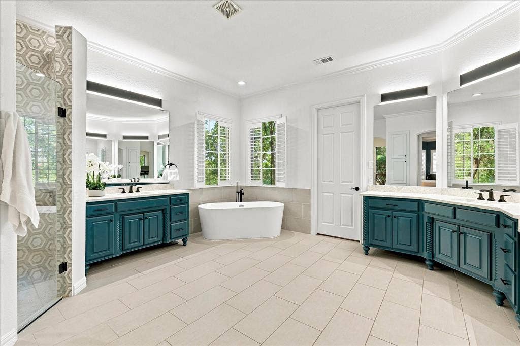 Dual vanities with integrated sinks, freestanding soaking tub, walk-in shower with hexagonal tile, crown molding, and plantation shutters