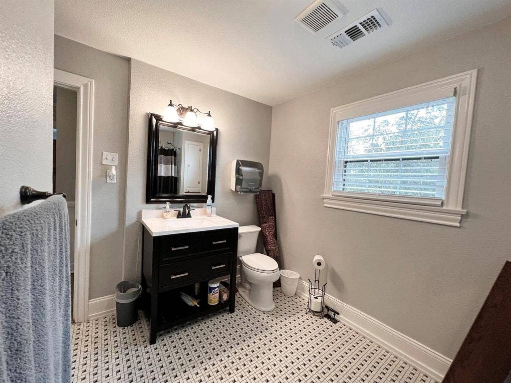 434 S Vine Avenue, Tyler, TX 75702 White and black patterned tile flooring, black vanity with white countertop, framed vanity mirror with sconce lighting, white window with blinds, and white toilet