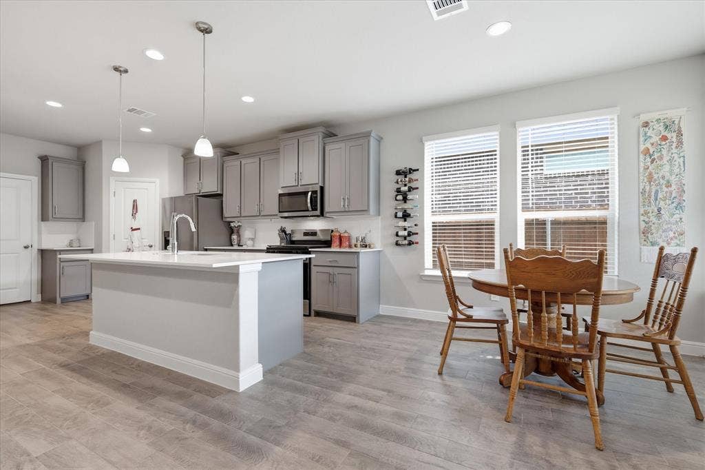 Open-concept kitchen and dining area featuring gray cabinetry, a central island with a sink, stainless steel appliances, and wood-finish flooring