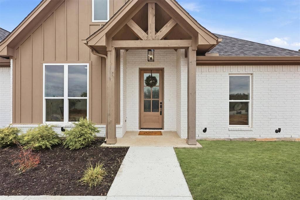 124 Norene Lane, Weatherford, TX 76088 Crisp white brick exterior with board and batten accents, a covered entry, wood-paneled front door, and a concrete walkway
