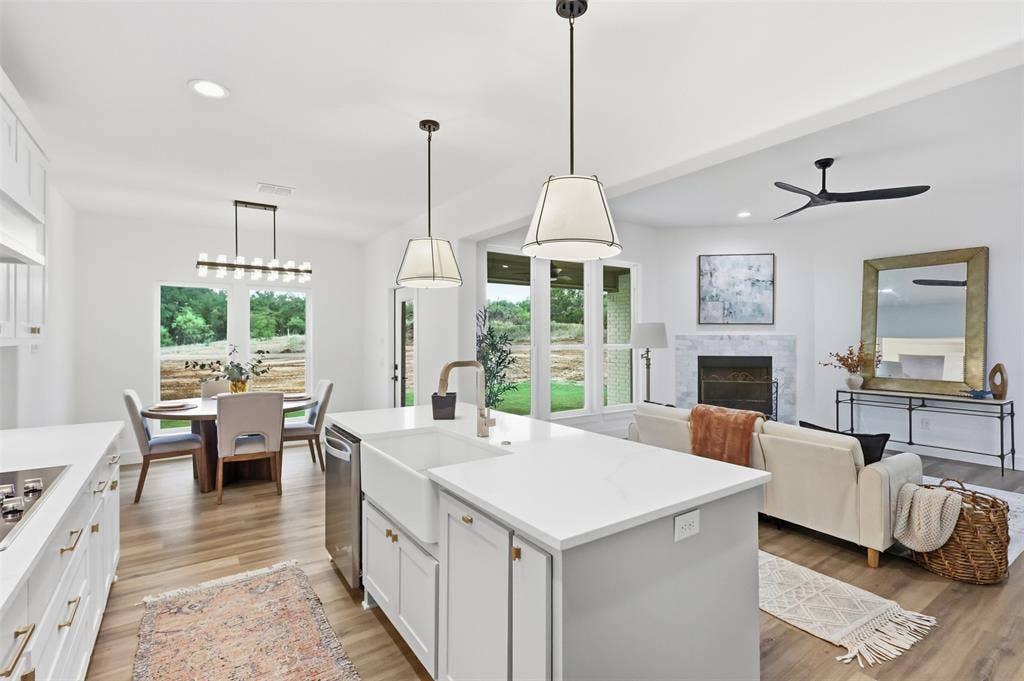 124 Norene Lane, Weatherford, TX 76088 Open-concept living space featuring wood-finish flooring, a kitchen island with a farmhouse sink, a dining area with a modern chandelier, a fireplace with a stone surround, and an integrated ceiling fan