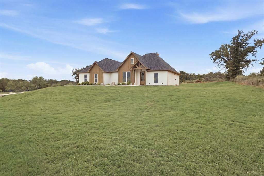 124 Norene Lane, Weatherford, TX 76088 Expansive green lawn surrounds a residence featuring a dark shingle roof, wood siding accents, and white exterior walls