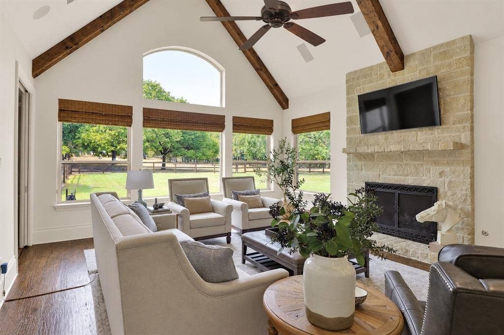 Vaulted ceiling with exposed wood beams, stone-clad fireplace with mantel, large arched window, multiple windows with woven shades, and wood-finish flooring