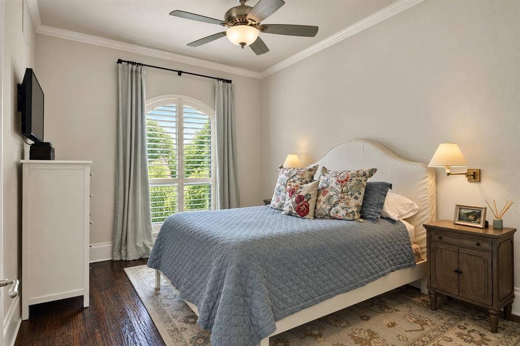 Arched window with plantation shutters, dark wood-finish flooring, ceiling fan with integrated lighting, crown molding, and wall-mounted sconces