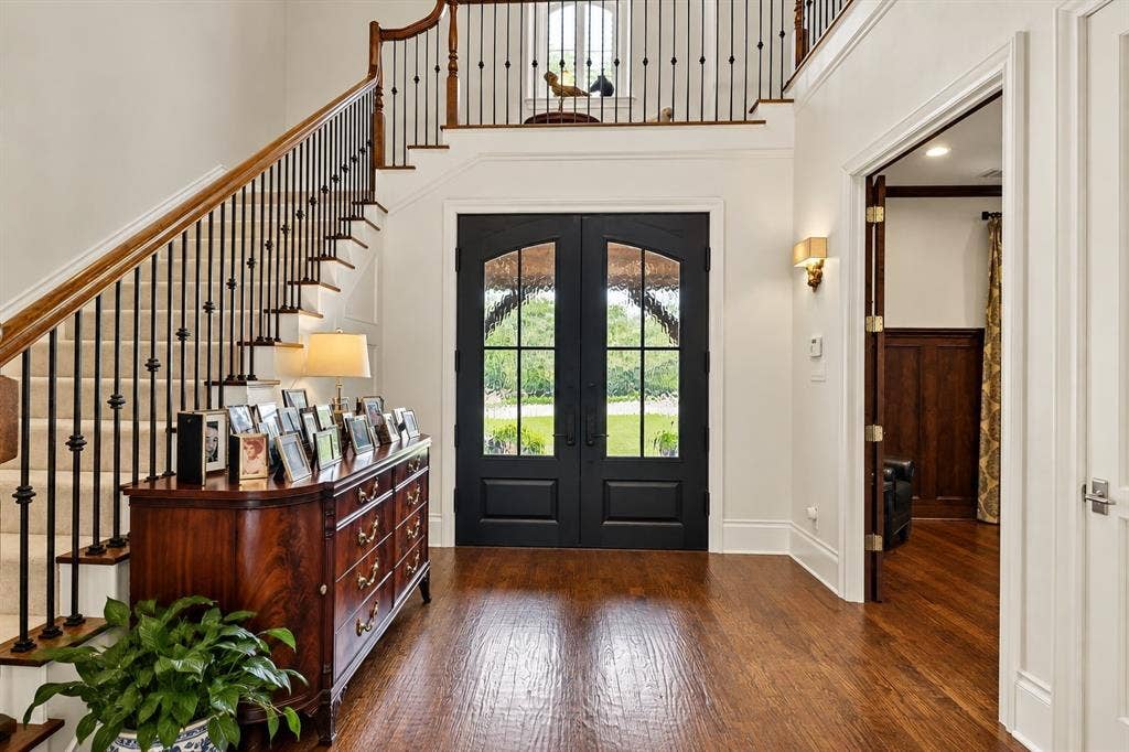 Grand entryway with wood-finish flooring, a carpeted staircase featuring wrought iron and wood railings, and double dark-paneled doors with glass inserts