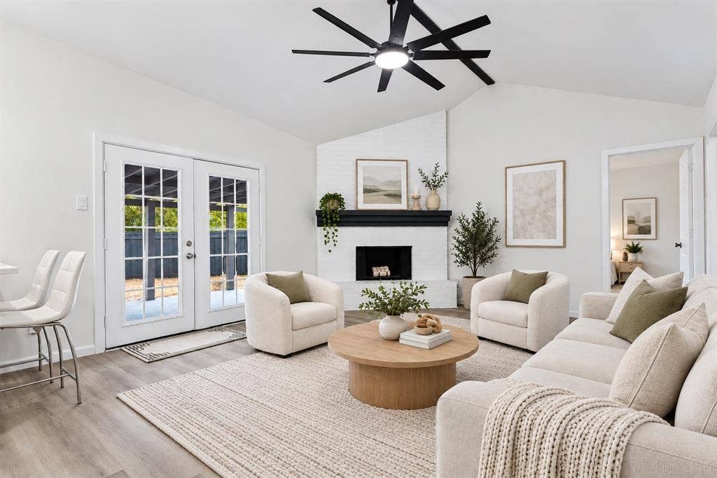 Vaulted ceilings, wood-finish flooring, white brick fireplace with a black mantel, and double French doors with grid-patterned glass