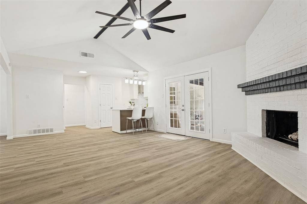 Vaulted ceiling with a contemporary ceiling fan, painted brick fireplace with a dark mantel, wood-finish flooring, and double French doors with grid-patterned glass