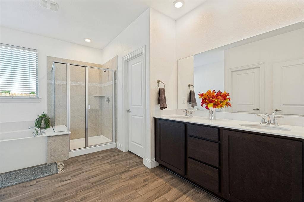Dual vanity with white countertops, wood-finish flooring, a tiled walk-in shower with glass enclosure, and a built-in soaking tub