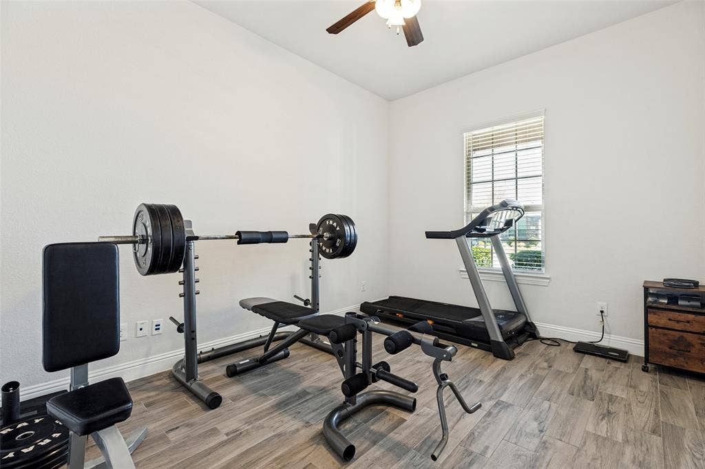 Recessed window with blinds, wood-finish flooring, ceiling fan with integrated lighting, and white walls. Currently used as a home gym