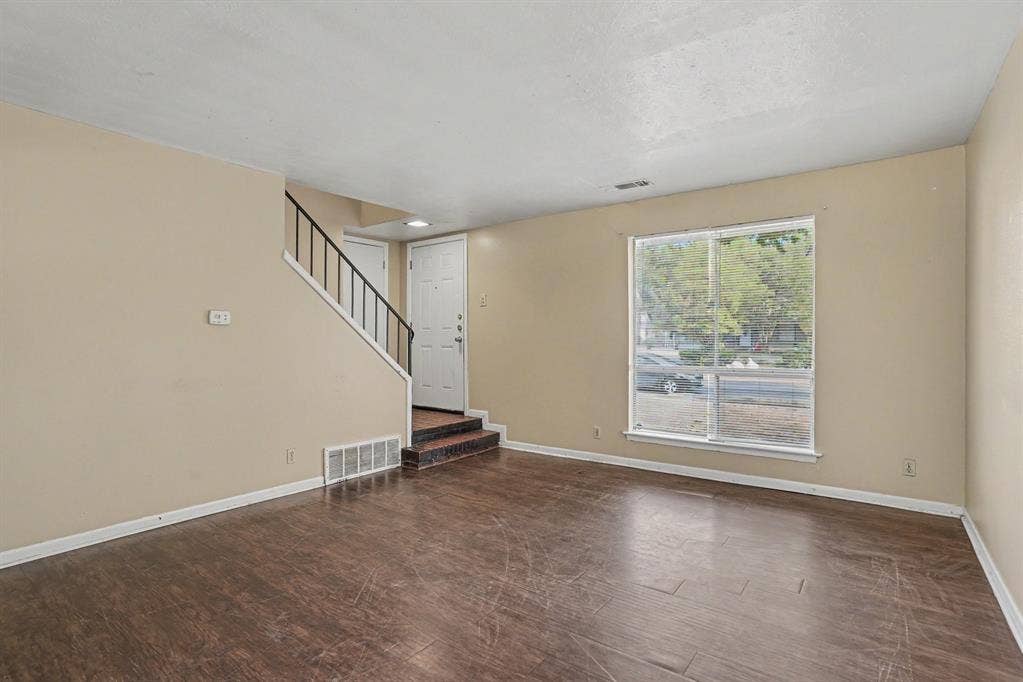 1414 Finley Road, Irving, TX 75062 Spacious living area featuring wood-finish flooring, a large window, neutral wall tones, and a staircase with a black metal railing