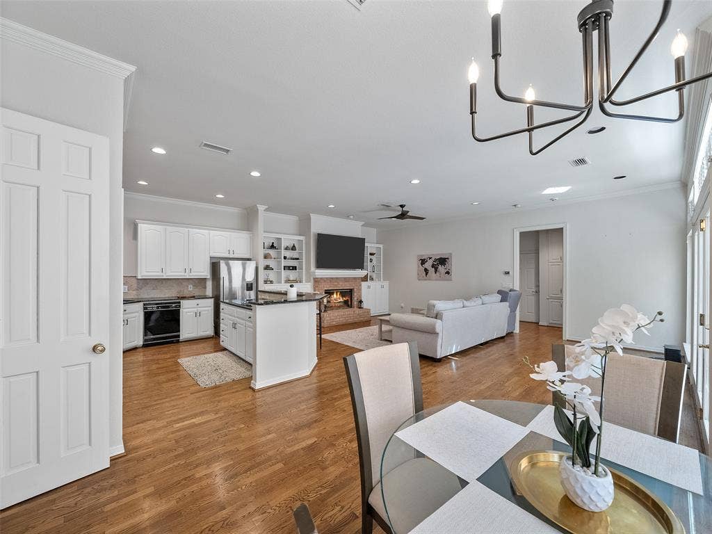 8640 Breakers Point, Dallas, TX 75243 Wood-finish flooring throughout, kitchen with white cabinetry and stainless steel appliances, brick fireplace with built-in shelving, high ceilings with recessed lighting, and a contemporary chandelier