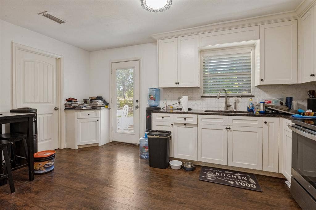 White shaker-style cabinetry with dark hardware, dark countertops, a tile backsplash, stainless steel sink with a gooseneck faucet, and wood-finish flooring