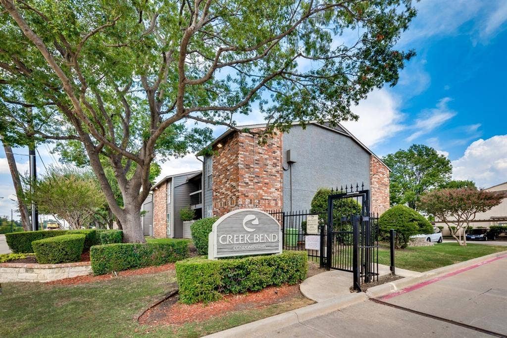 Brick and stucco exterior with lush landscaping, mature tree, and a decorative wrought iron gate