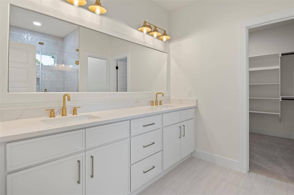 Dual vanity with gold-finish faucets and hardware, white cabinetry, a full-width mirror, and tile flooring