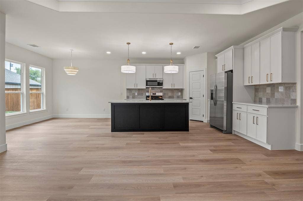 Spacious kitchen and living area featuring wood-finish flooring, white cabinetry with gold-tone hardware, a black kitchen island, stainless steel appliances, and ample natural light from multiple windows