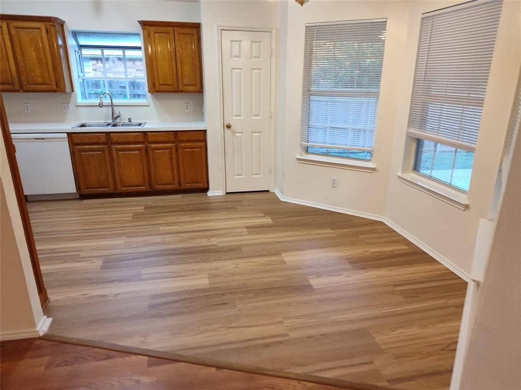 Kitchen featuring wood-finish cabinetry, hard-surface countertops, a stainless steel sink with a gooseneck faucet, light-toned wood-finish flooring, and multiple windows with horizontal blinds