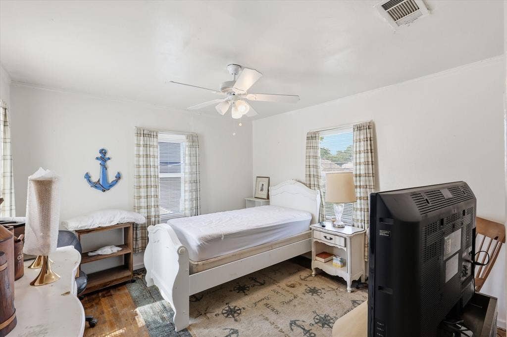 6807 Prosper Street, Dallas, TX 75209 Light-filled bedroom with white walls, wood flooring, ceiling fan, multiple windows, and a flexible layout. The room offers space for a bed, nightstand, dresser, seating, or media setup, with natural light creating a bright and comfortable atmosphere. Soft curtains and simple finishes make this room easy to personalize as a restful retreat, guest space, or home office.