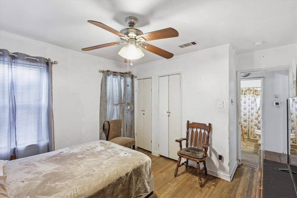 6807 Prosper Street, Dallas, TX 75209 Bedroom view highlighting the ceiling fan, wood flooring, closet doors, multiple windows, and direct sightline toward the bathroom. The room provides functional storage, natural light, and enough space for bedroom furniture plus a small seating area. With white walls and warm floors, this space offers a clean, flexible canvas ready for everyday use or future personal updates.
