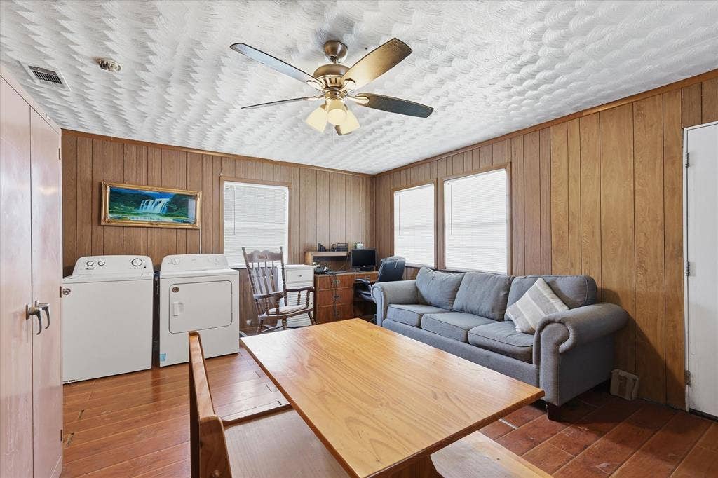 6807 Prosper Street, Dallas, TX 75209 Living room view highlighting the ceiling fan, textured ceiling detail, wood-look floors, paneled walls, and flexible furniture arrangement. With natural light from multiple windows and space for seating, laundry, and work-from-home use, this room offers practical everyday versatility.