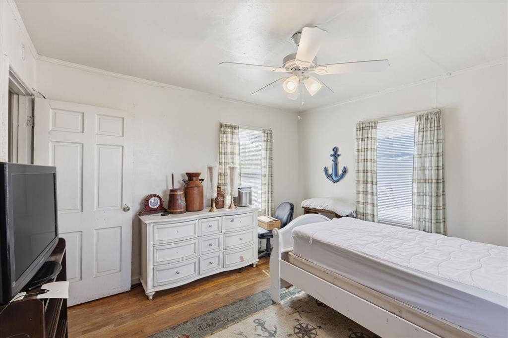 6807 Prosper Street, Dallas, TX 75209 Bedroom view featuring wood flooring, white walls, ceiling fan, closet door, dresser space, and two windows with patterned curtains. The room feels bright and functional, with enough wall space for furniture placement and personal décor. Its practical size and natural light make it well suited for a bedroom, guest room, office, hobby space, or other flexible use.