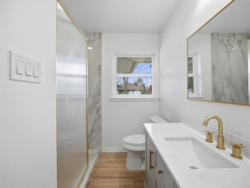 10270 Kilkenny Place, Dallas, TX 75228 Walk-in shower with opaque glass paneling, wood-finish flooring, single vanity with integrated sink, rectangular gold-framed mirror, and gold-finish fixtures