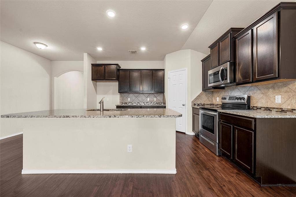 3217 Beutel Road, Lorena, TX 76655 Kitchen with appliances with stainless steel finishes, light stone counters, dark brown cabinets, an island with sink, and dark wood-style floors