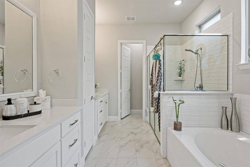 Spacious bathroom featuring a built-in soaking tub, glass-enclosed shower, dual vanities with white cabinetry, and marble-look tile flooring