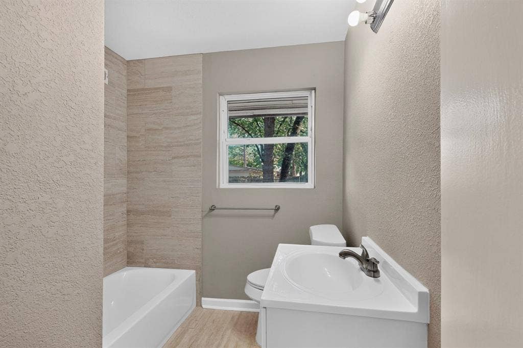 2314 Wales Place, Irving, TX 75062 Bathroom featuring a white vanity with integrated sink, a toilet, a window, and a bathtub with tiled surround