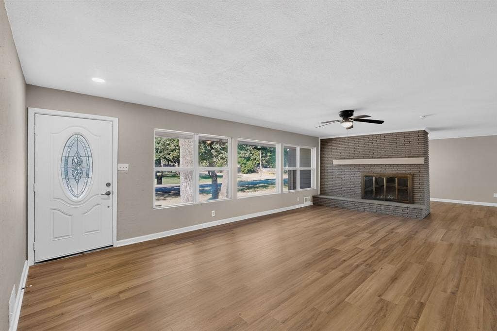 2314 Wales Place, Irving, TX 75062 Spacious living area featuring wood-finish flooring, a brick fireplace with a mantle, a ceiling fan with integrated lighting, and multiple windows
