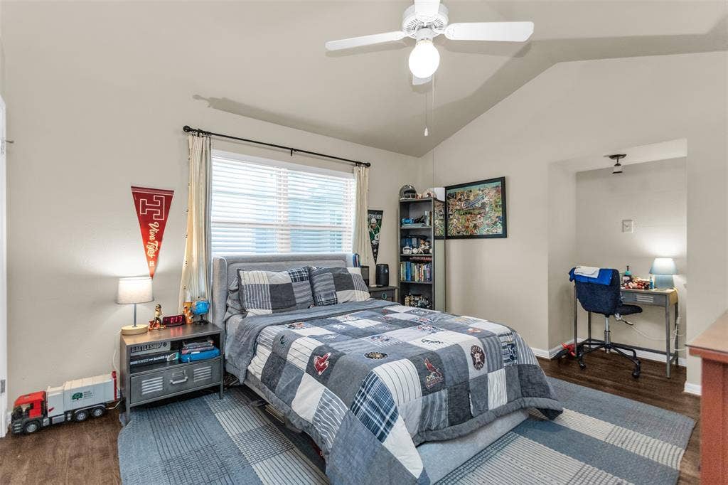 Spacious bedroom featuring vaulted ceiling, ceiling fan with integrated lighting, large windows with plantation shutters, wood-finish flooring, and a coordinating area rug