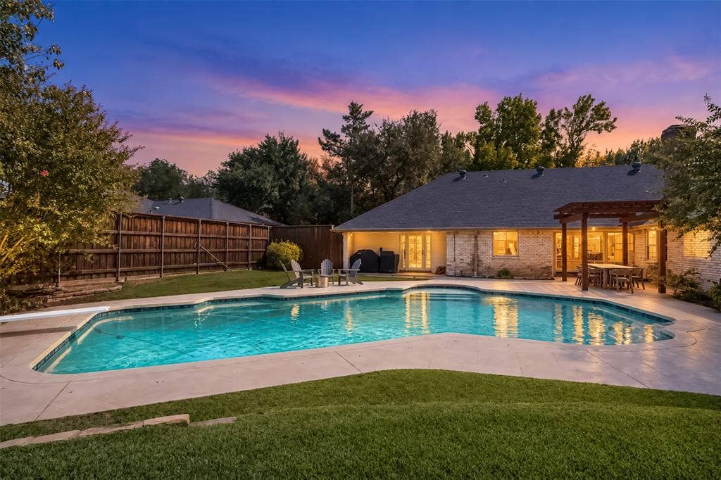 Resort-style backyard oasis featuring a sparkling resort-pool with curved edges and integrated lighting, expansive stamped concrete decking, manicured lawn, mature landscaping with mixed tree canopy, and a contemporary single-story residence with illuminated covered patio areas and multiple sets of glass doors opening to the outdoor living space