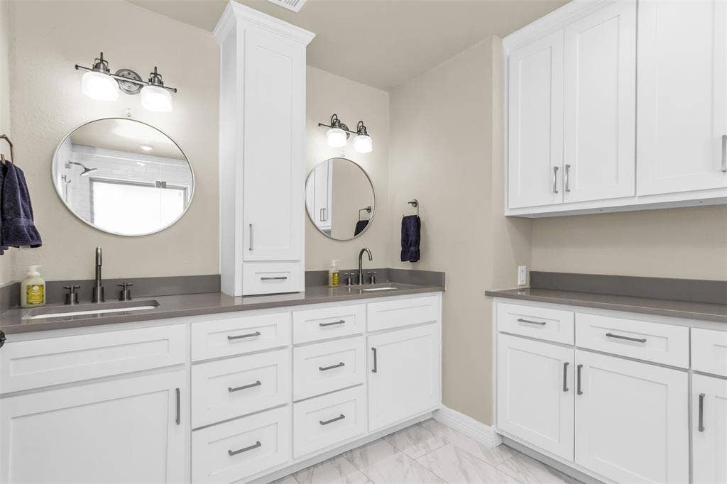 Dual-vanity primary bathroom featuring white shaker-style cabinetry, gray quartzite countertops, polished round mirrors.
