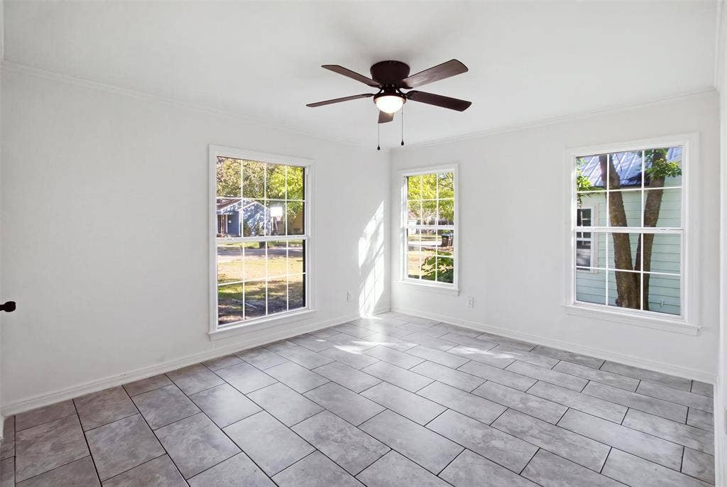 1105 Bell Drive, Greenville, TX 75401 Light-filled room featuring three large windows, a ceiling fan with integrated lighting, crown molding, and large format tile flooring