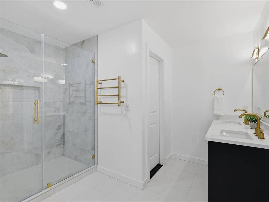 2500 Teakwood Lane, Plano, TX 75075 Spacious bathroom featuring a glass-enclosed shower with stone-finish tile, a dual vanity with a white countertop and gold-finish fixtures, and gold-finish lighting