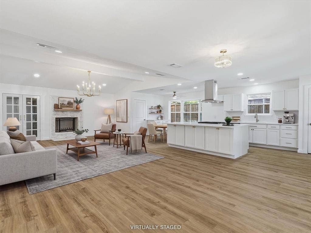 2500 Teakwood Lane, Plano, TX 75075 Open-concept living space featuring wood-finish flooring, a white kitchen island with counter seating, a stainless steel range hood, a fireplace with a white brick surround, and white kitchen cabinetry