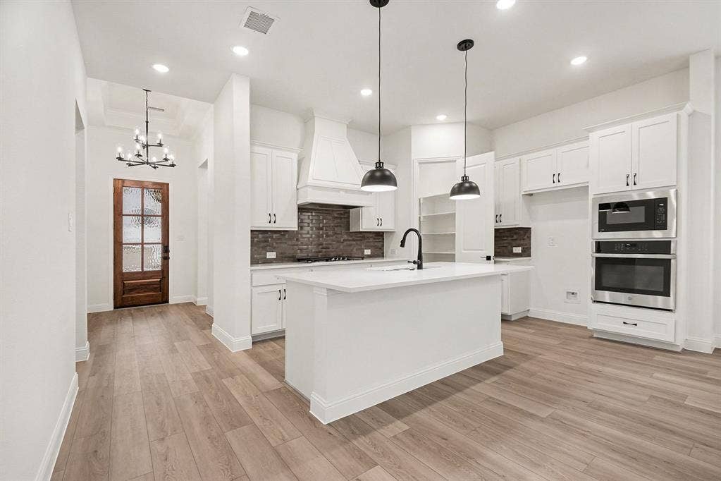 Kitchen featuring white shaker-style cabinetry, central island with undermount sink, brick-style backsplash, integrated double wall ovens, and wood-finish flooring