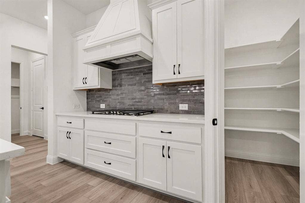 Kitchen featuring white cabinetry, a built-in range hood, a tiled backsplash, and a walk-in pantry with wrap-around shelving